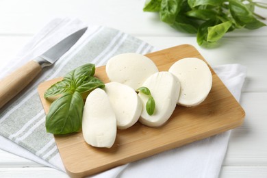 Photo of Pieces of delicious mozzarella cheese, basil leaves and knife on white wooden table, closeup