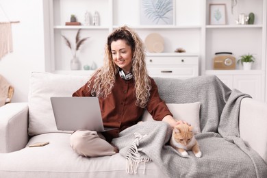 Photo of Smiling woman working with laptop and stroking cat on sofa in home office