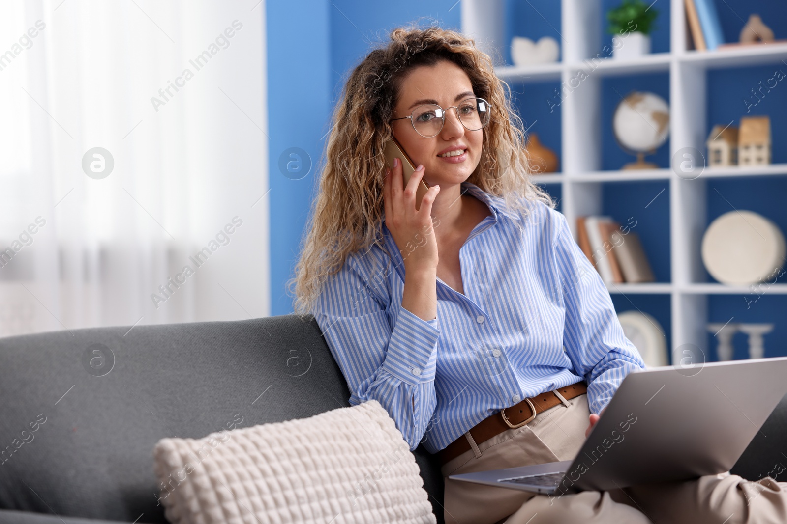 Smiling woman with laptop talking by smartphone on sofa in home office Photo of Smiling woman with laptop talking by smartphone on sofa in home office