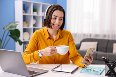 Photo of Smiling woman with smartphone and cup of hot drink at desk in home office