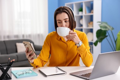 Photo of Woman with smartphone and cup of hot drink at desk in home office