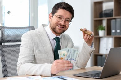 Photo of Happy greedy banker with dollar banknotes and cigar at table in office