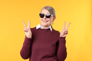 Photo of Happy senior woman in stylish outfit showing V-signs on orange background