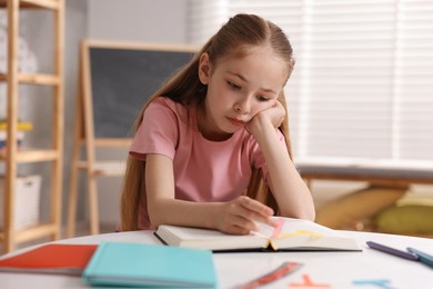 Photo of Dyslexia. Upset little girl reading book at table indoors