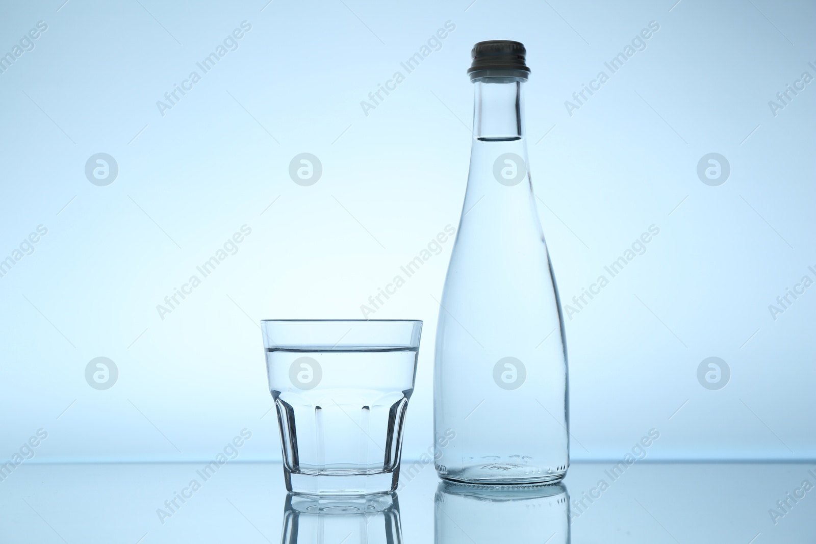 Bottle and glass of water on mirror table against light blue gradient background Photo of Bottle and glass of water on mirror table against light blue gradient background