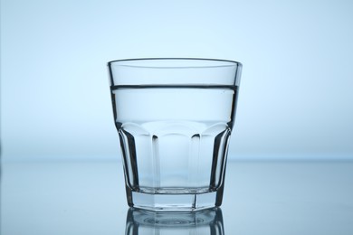 Photo of Fresh water in glass on mirror table against light blue gradient background, closeup