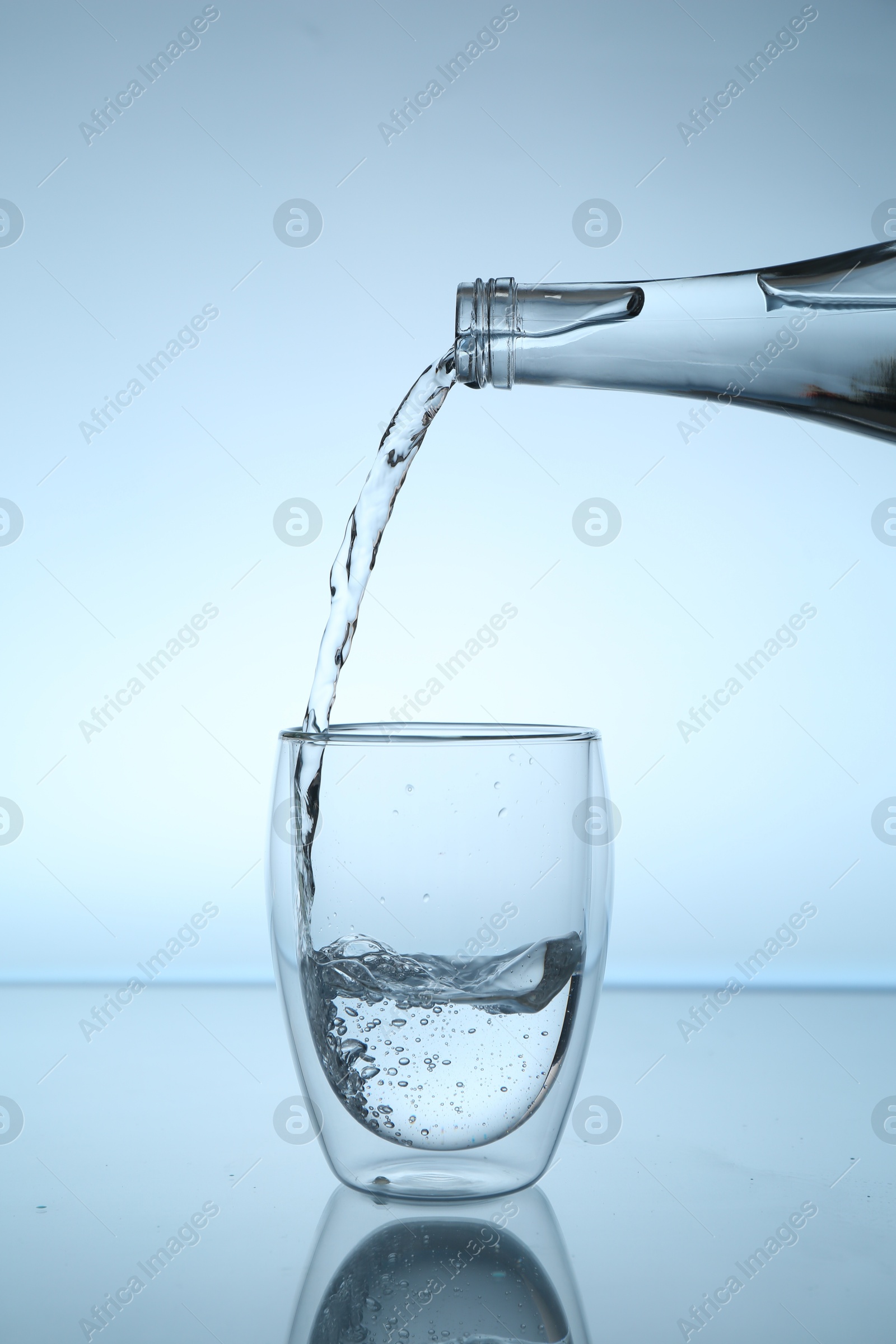 Pouring water from bottle into glass at mirror table against light blue gradient background, closeup Photo of Pouring water from bottle into glass at mirror table against light blue gradient background, closeup