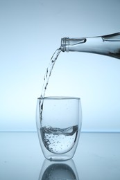 Photo of Pouring water from bottle into glass at mirror table against light blue gradient background, closeup