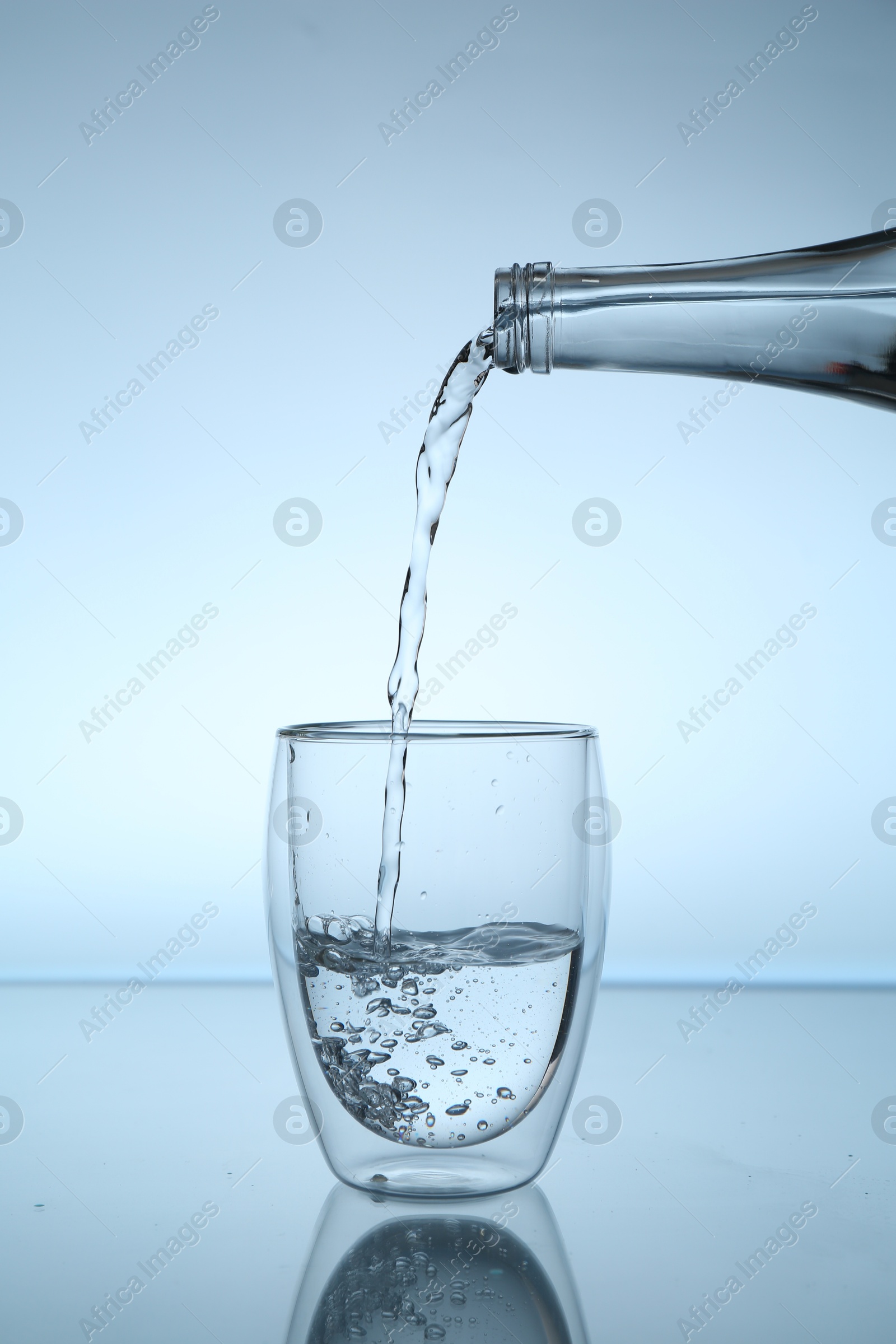 Pouring water from bottle into glass at mirror table against light blue gradient background, closeup Photo of Pouring water from bottle into glass at mirror table against light blue gradient background, closeup