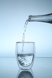 Photo of Pouring water from bottle into glass at mirror table against light blue gradient background, closeup