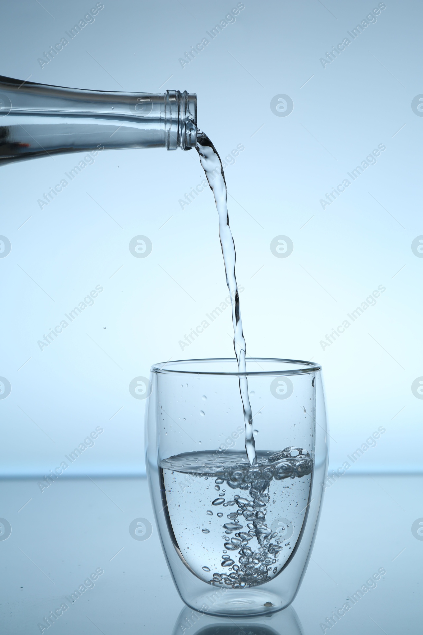 Photo of Pouring water from bottle into glass at mirror table against light blue gradient background, closeup