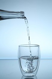 Photo of Pouring water from bottle into glass at mirror table against light blue gradient background, closeup