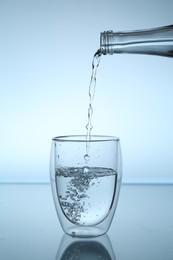 Photo of Pouring water from bottle into glass at mirror table against light blue gradient background, closeup