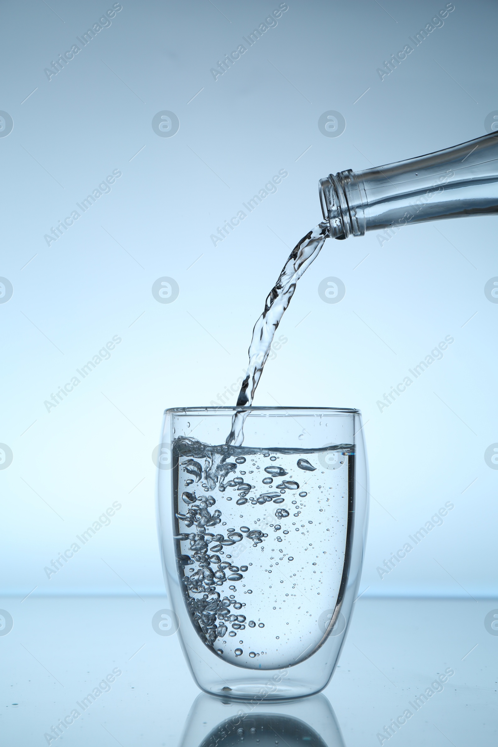 Photo of Pouring water from bottle into glass at mirror table against light blue gradient background, closeup