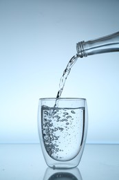 Photo of Pouring water from bottle into glass at mirror table against light blue gradient background, closeup