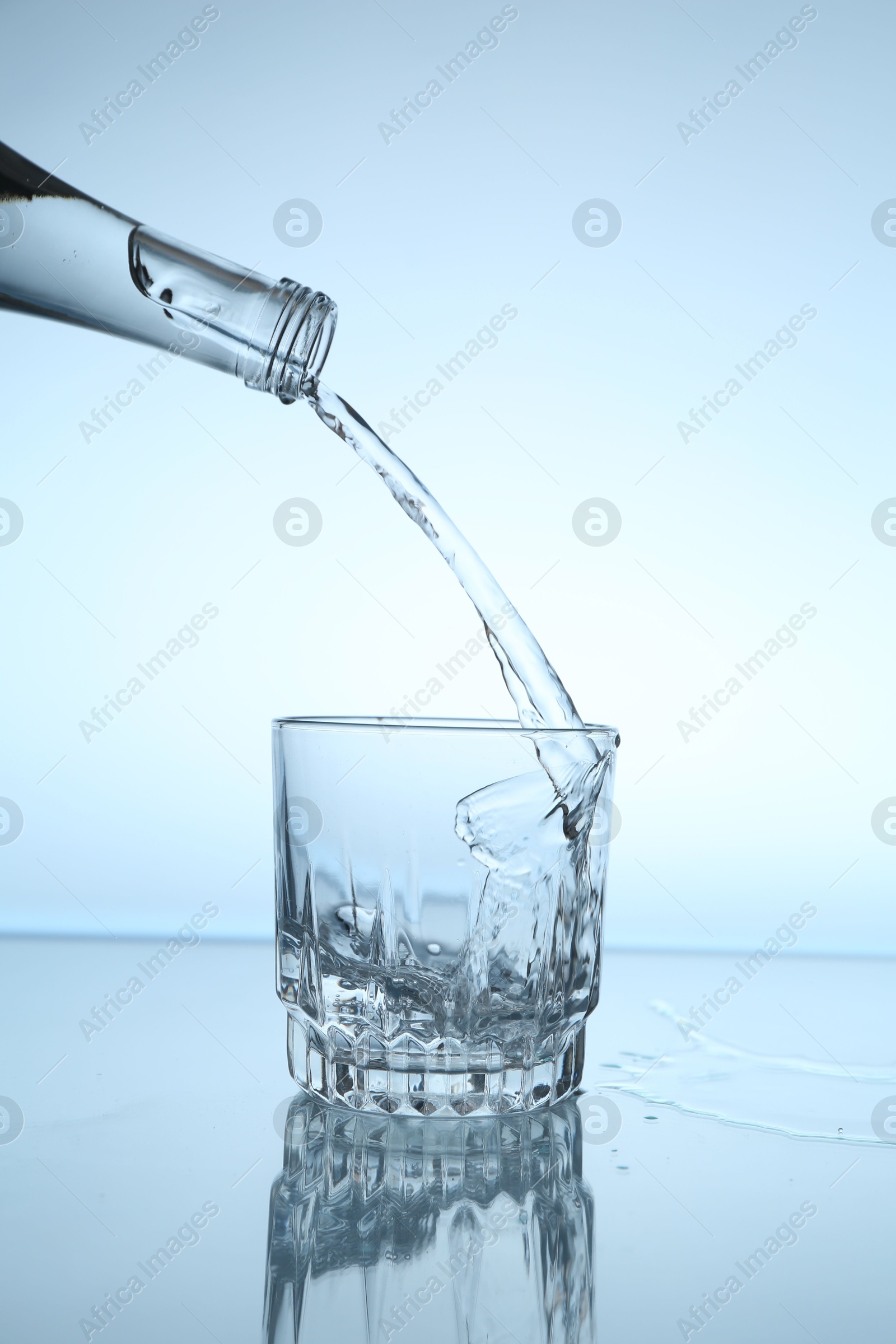 Photo of Pouring water from bottle into glass at mirror table against light blue gradient background, closeup
