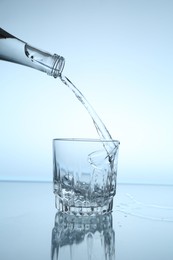 Photo of Pouring water from bottle into glass at mirror table against light blue gradient background, closeup