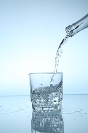 Photo of Pouring water from bottle into glass at mirror table against light blue gradient background, closeup