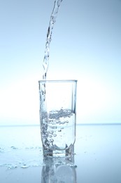 Photo of Pouring water into glass at mirror table against light blue gradient background, closeup