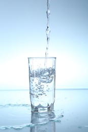 Photo of Pouring water into glass at mirror table against light blue gradient background, closeup
