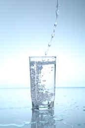 Photo of Pouring water into glass at mirror table against light blue gradient background, closeup