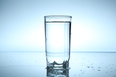 Photo of Fresh water in glass on mirror table against light blue gradient background, closeup