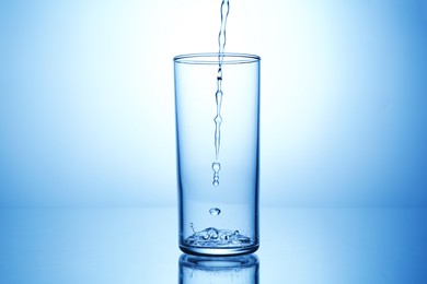 Photo of Pouring water into glass at mirror table against light blue gradient background, closeup