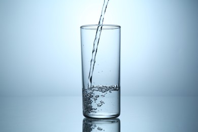 Photo of Pouring water into glass at mirror table against light grey gradient background, closeup
