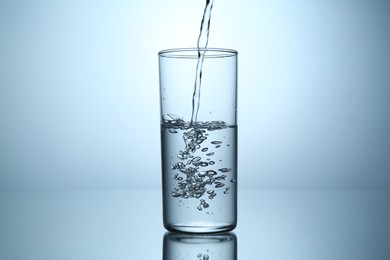 Photo of Pouring water into glass at mirror table against light grey gradient background, closeup