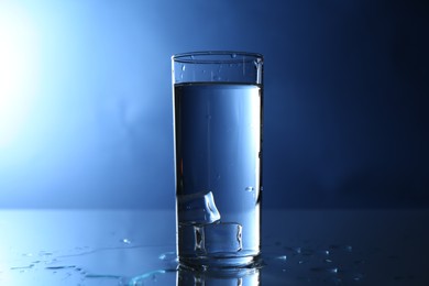 Photo of Fresh water with ice cubes in glass on mirror table against blue gradient background, closeup