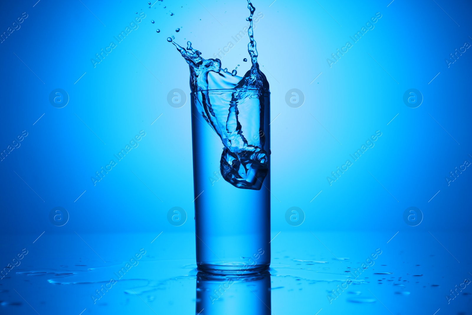Fresh water with ice cube splashing out of glass on mirror table against blue gradient background Photo of Fresh water with ice cube splashing out of glass on mirror table against blue gradient background
