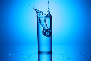 Photo of Fresh water with ice cube splashing out of glass on mirror table against blue gradient background