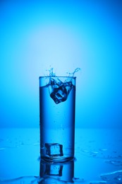 Photo of Fresh water with ice cubes splashing out of glass on mirror table against blue gradient background