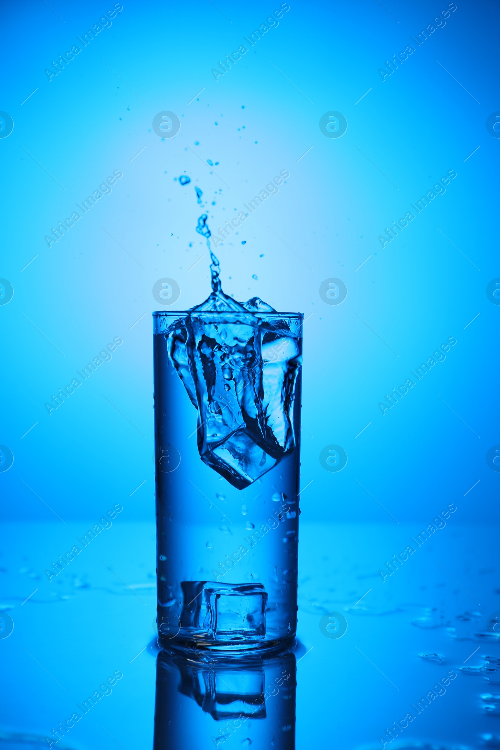 Fresh water with ice cubes splashing out of glass on mirror table against blue gradient background Photo of Fresh water with ice cubes splashing out of glass on mirror table against blue gradient background