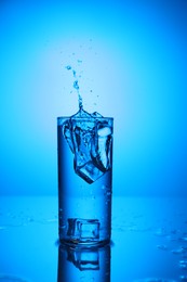 Photo of Fresh water with ice cubes splashing out of glass on mirror table against blue gradient background