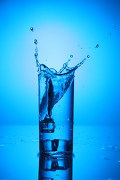 Photo of Fresh water with ice cubes splashing out of glass on mirror table against blue gradient background