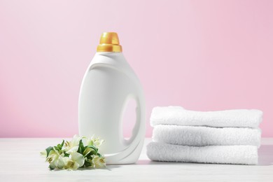 Photo of Bottle of fabric softener, terry towels and lily flowers on white wooden table against pink background