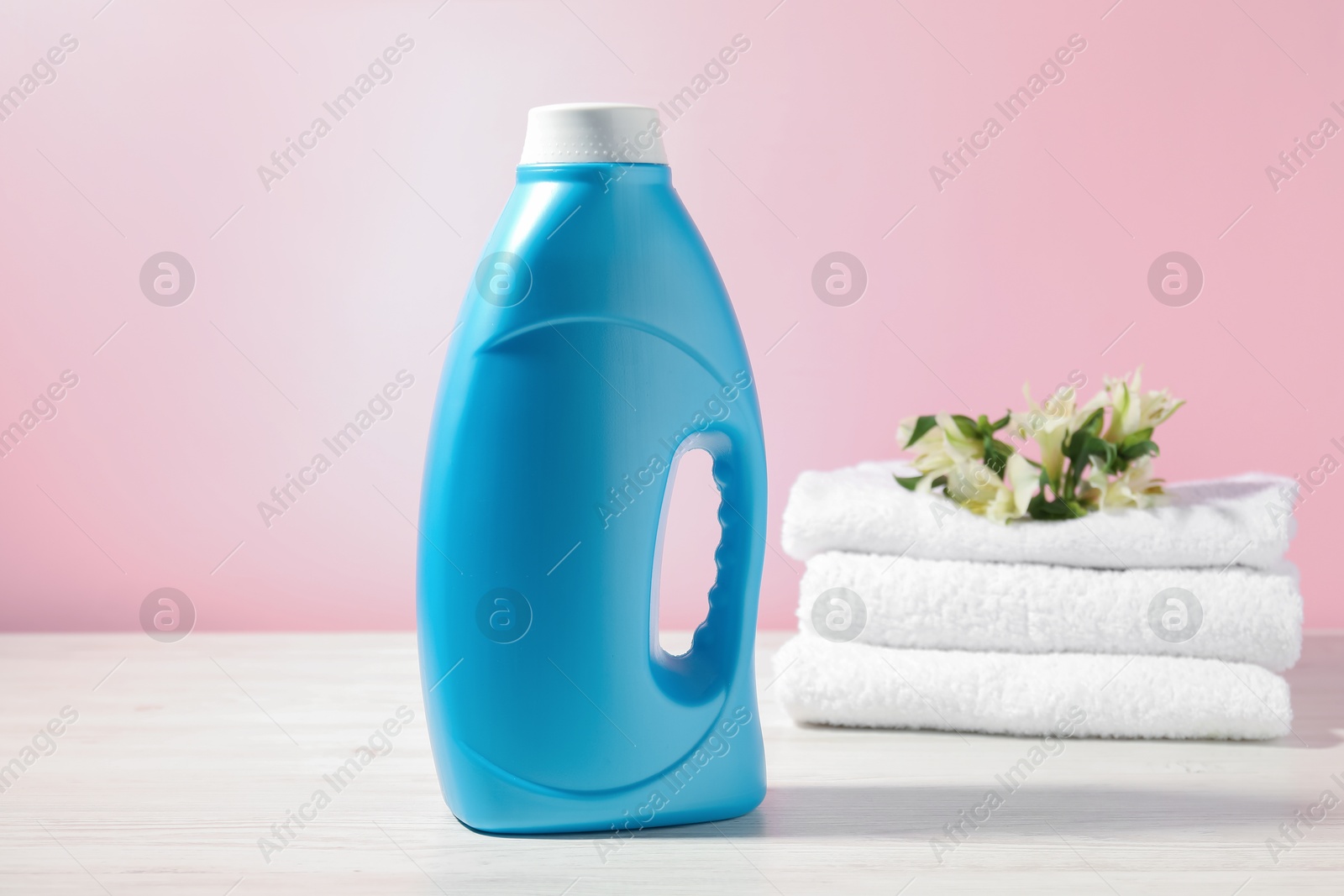 Bottle of fabric softener, terry towels and lily flowers on white wooden table against pink background Photo of Bottle of fabric softener, terry towels and lily flowers on white wooden table against pink background