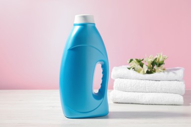 Photo of Bottle of fabric softener, terry towels and lily flowers on white wooden table against pink background