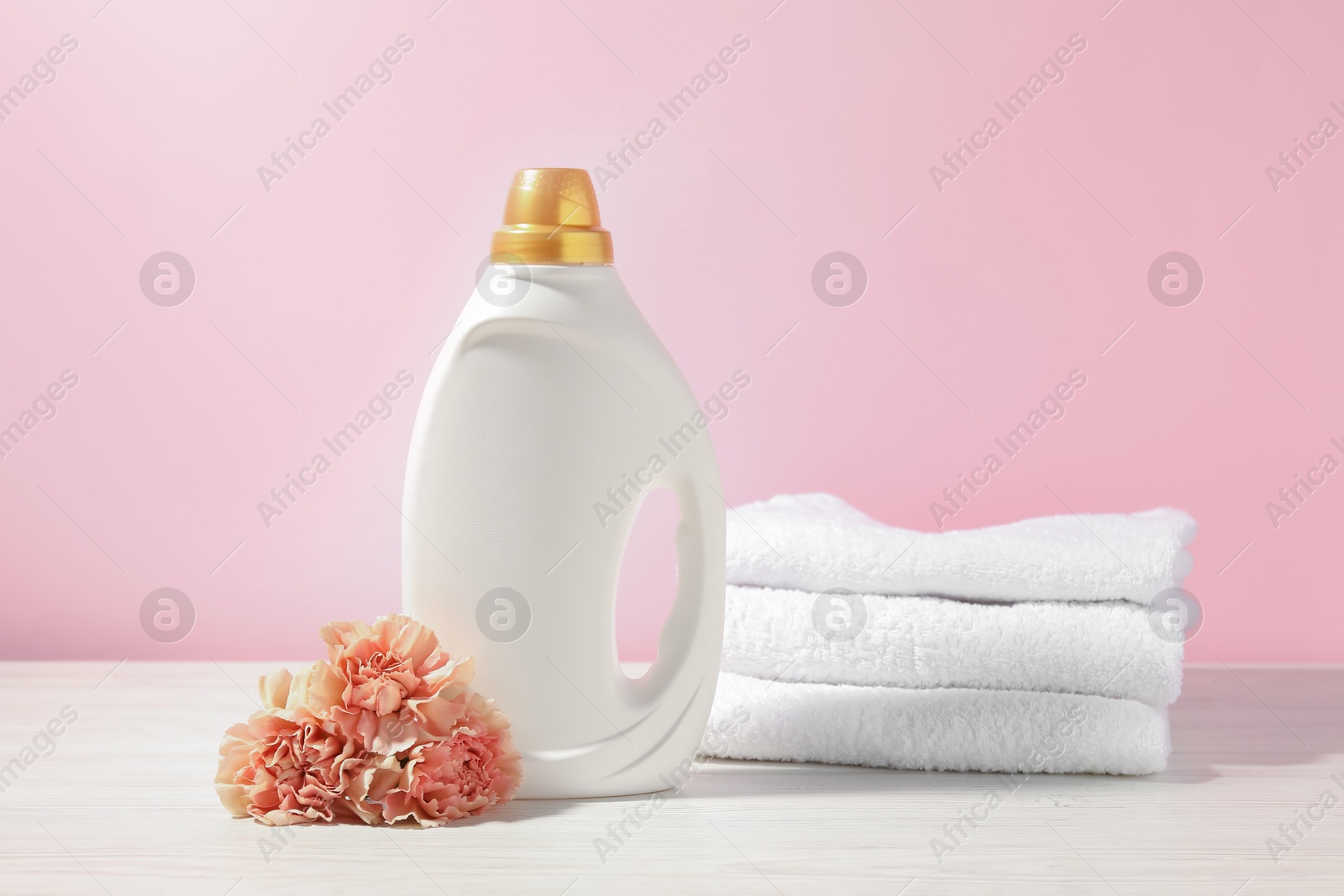 Bottle of fabric softener, terry towels and carnation flowers on white wooden table against pink background Photo of Bottle of fabric softener, terry towels and carnation flowers on white wooden table against pink background