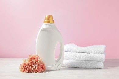 Photo of Bottle of fabric softener, terry towels and carnation flowers on white wooden table against pink background