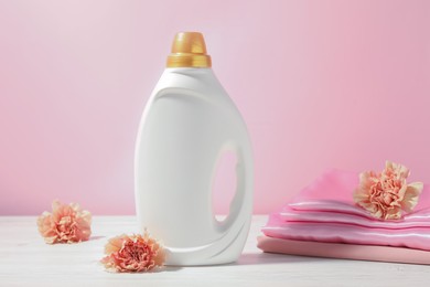 Photo of Bottle of fabric softener, clean clothes and carnation flowers on white wooden table against pink background
