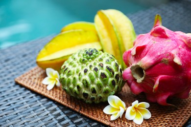 Photo of Different tropical fruit on wicker table near swimming pool, closeup