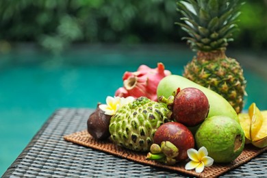 Photo of Different tropical fruit on wicker table near swimming pool. Space for text