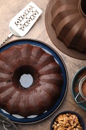 Photo of Tasty chocolate bundt cake, walnuts, cocoa powder and server on wooden table, flat lay
