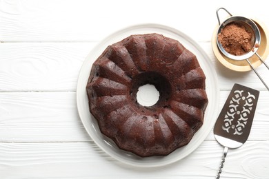 Photo of Tasty chocolate bundt cake, cocoa powder and server on white wooden table, flat lay. Space for text