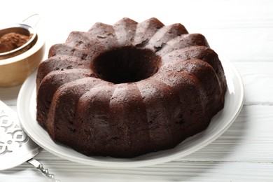 Photo of Tasty chocolate bundt cake, cocoa powder and server on white wooden table, closeup
