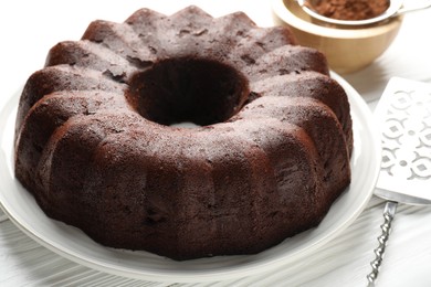 Photo of Tasty chocolate bundt cake, cocoa powder and server on white wooden table, closeup