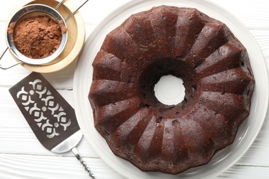 Photo of Tasty chocolate bundt cake, cocoa powder and server on white wooden table, flat lay