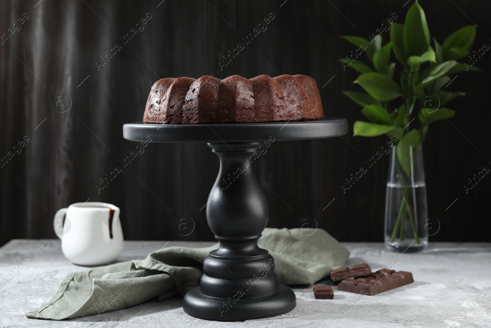 Tasty bundt cake, pieces of chocolate, sauce and green leaves on grey table Photo of Tasty bundt cake, pieces of chocolate, sauce and green leaves on grey table